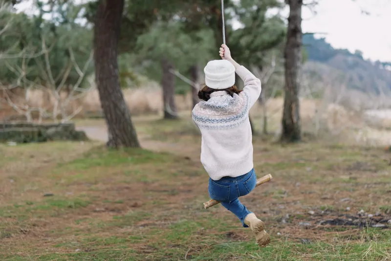 Person on a swing in an outdoor park, wearing a cozy sweater, jeans and a hat, captured mid motion among trees and open grass.