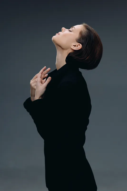 Elegant woman in black coat poses with closed eyes against gray background, displaying calm and grace in a minimalist style.