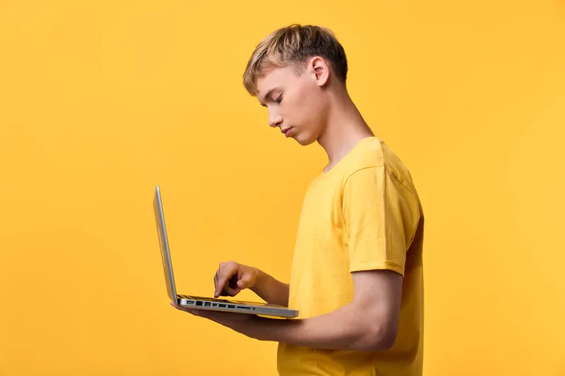 Laptop held by a focused man in a yellow tee, side view against a bright backdrop, highlighting technology use, online work, and casual learning in a modern workspace.