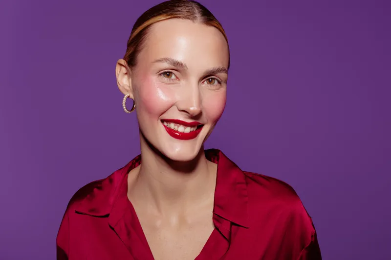 Smiling woman with red lipstick and shiny skin wears red blouse and hoop earrings. Portrait on purple background with soft lighting. Concept of beauty, fashion, and confident expression.