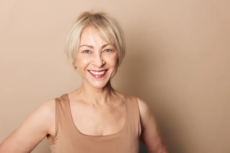 Smiling mature woman with short blonde hair wears beige tank top, posing confidently against neutral background. Concept of aging beauty, happiness, wellness, and natural lifestyle.