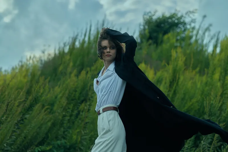 Fashion poised in a white dress with a flowing black cape, the model stands in tall grass as wind sweeps the scene, creating dramatic movement and a cinematic outdoor look.