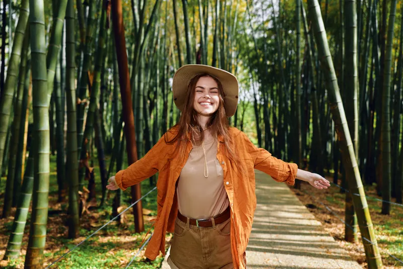 Woman in orange jacket and hat smiling with arms out on a bamboo forest path, casual outdoor portrait with sunlight, travel and nature mood, carefree autumn tones and green scenery.