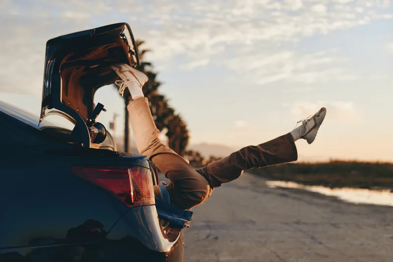 Woman lifestyle on road beside car at sunset, hands on car hood, relaxed travel stop, peaceful outdoor scene, wide sky and horizon, rural highway atmosphere, evening light