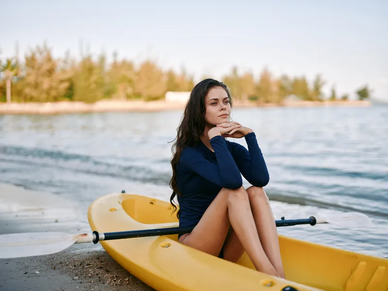 Young woman in a navy sweater sits in a yellow kayak on a quiet beach, gazing thoughtfully toward the sea as gentle waves lap the shore during golden hour.