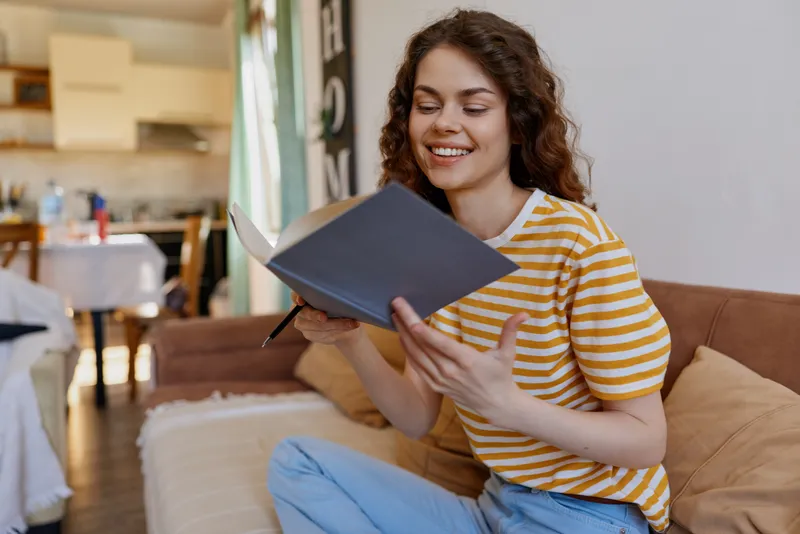 Woman indoors reading notebook, smiling, relaxed home moment, striped shirt, soft lighting, cozy sofa, casual lifestyle, warm colors, natural mood