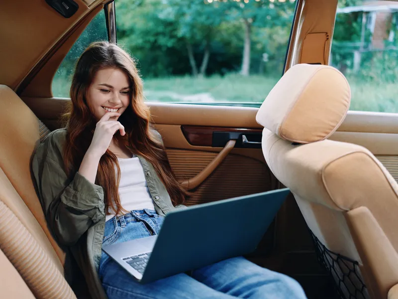Relaxed woman working on a laptop in a vintage car, enjoying her time with a smile, surrounded by greenery and sunlight. Stylish casual outfit enhances the atmosphere.