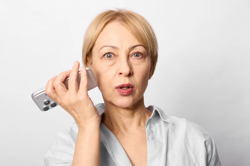 Woman elderly smartphone phone calling talking communication portrait closeup indoors, older woman holds smartphone to her ear, surprised worried expression, studio white background