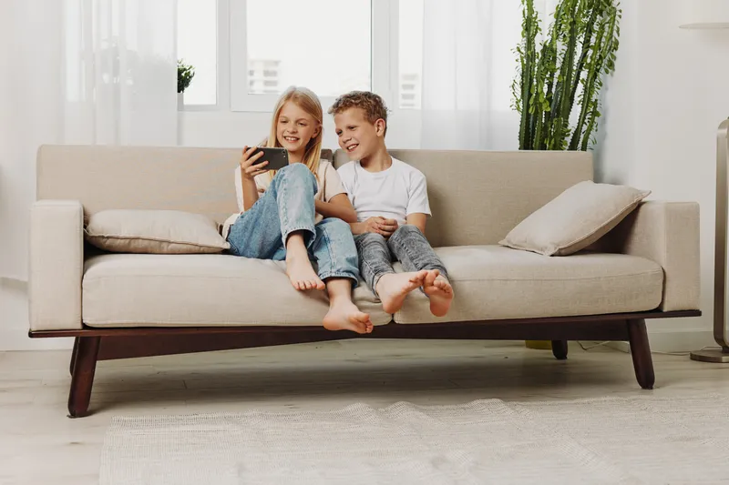 Two happy children sitting barefoot on beige sofa in bright living room, smiling and watching smartphone together. Concept of family, childhood, technology, entertainment, and modern lifestyle.