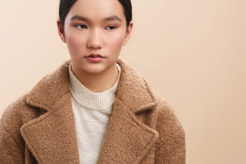 asian woman in a warm woolen coat stands against a soft beige backdrop, looking slightly to the side with a calm, poised expression that conveys subtle confidence and style.
