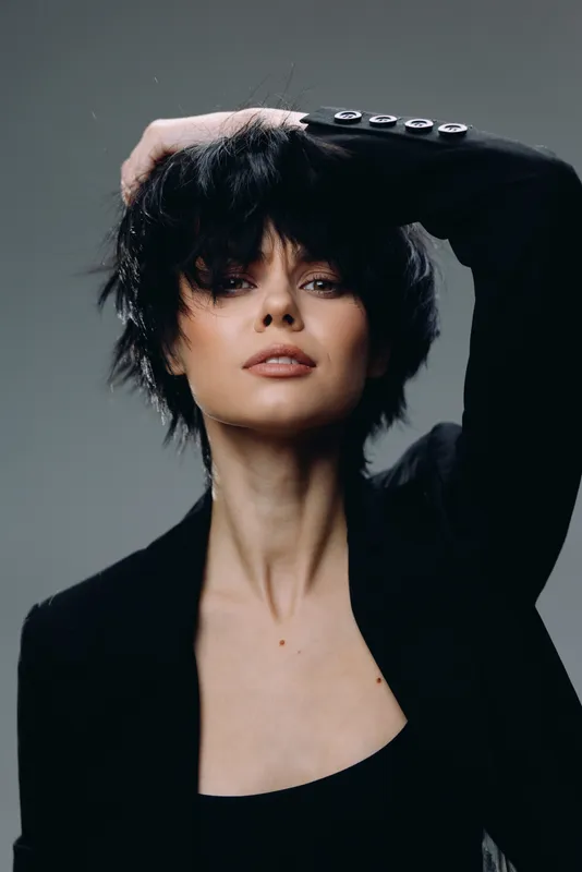 Stylish woman with short black hair posing confidently in dark clothing with a neutral gray background and soft lighting