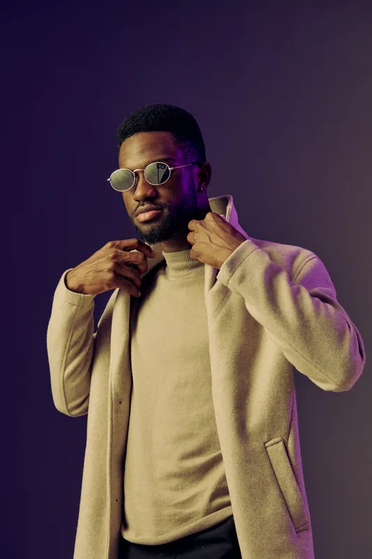Stylish African man wearing sunglasses and a beige coat against a purple backdrop, exuding confidence and modern fashion in his casual outfit