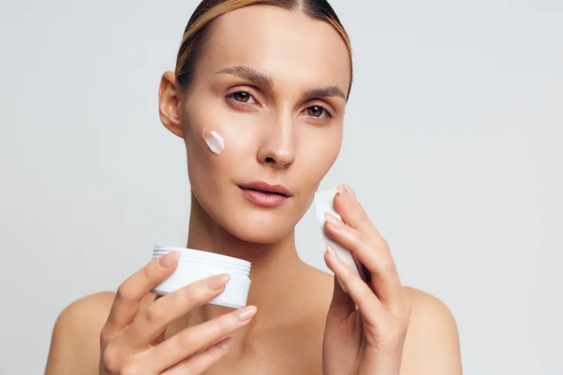 Young woman applying white face cream on cheek with fingers, holding jar. Close-up portrait on light background. Concept of skincare, beauty routine, and self-care with natural lighting.
