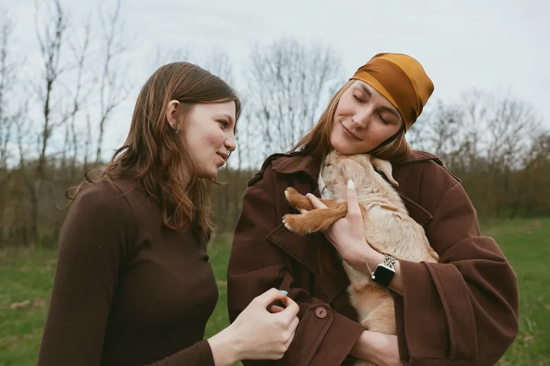 Two women outdoors with a gentle goat kid, sharing a warm moment, affection and friendship as the animal nuzzles them and the countryside air feels fresh