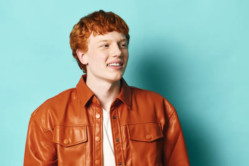 Young man with red hair wearing orange jacket smiling and looking away standing against plain light blue background. Portrait of casual style confident male in studio.