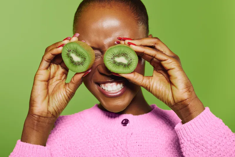 Happy dark skinned African woman in pink sweater holds two kiwi halves in front of her eyes, smiling with closed eyes. Bright green background and fresh fruit concept for health and joy.
