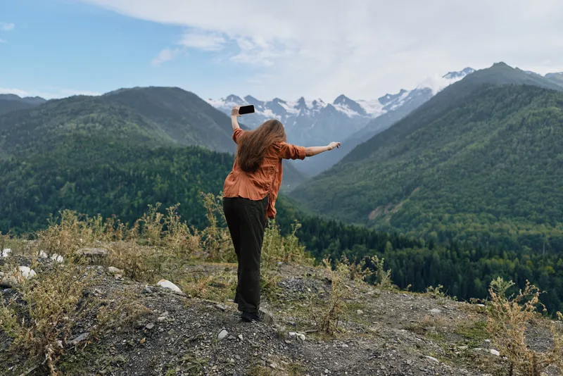 Traveler stands on a rocky overlook with arms raised, capturing a moment of freedom amid vast mountains and valley, wearing an orange jacket, embracing adventure and awe