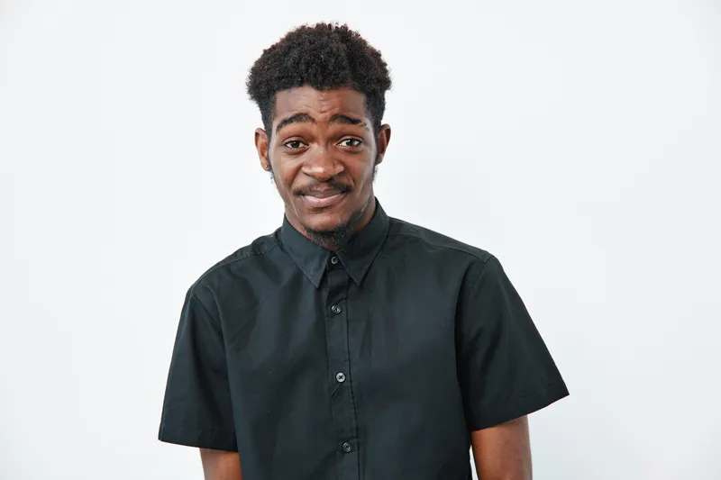 Young black man with dark skin wearing black shirt standing on white background looking at camera with uncertain expression and slight smile.