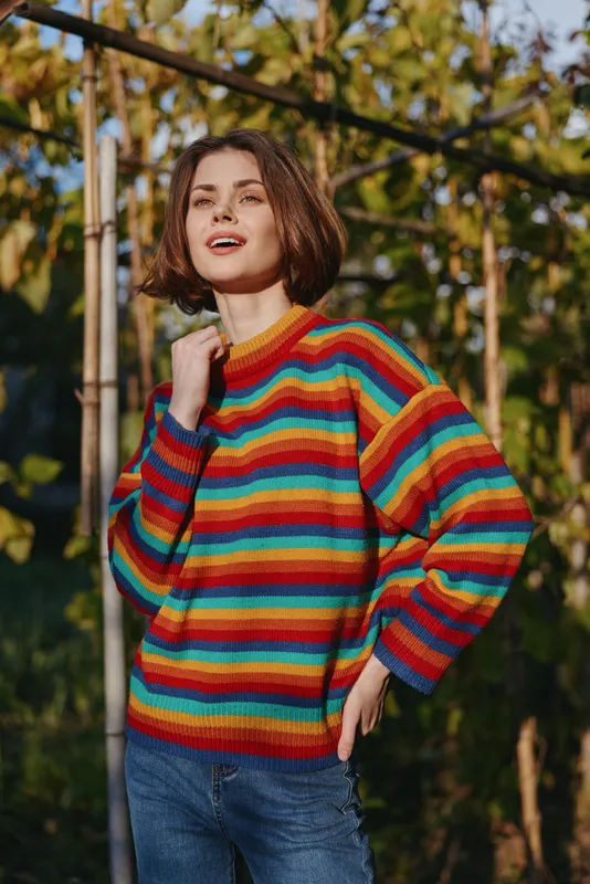 Woman in her early twenties with short brown hair smiling in a rainbow sweater and casual outfit, outdoor portrait in a garden background with warm sunlight, relaxed pose.