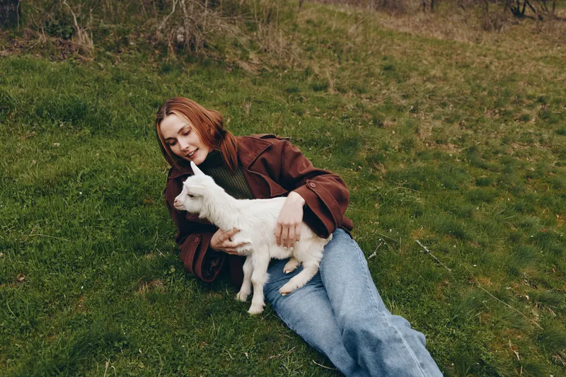 Young woman in a brown jacket lies on grass holding a white baby goat while smiling in a natural outdoor setting, capturing a peaceful lifestyle moment with animals.
