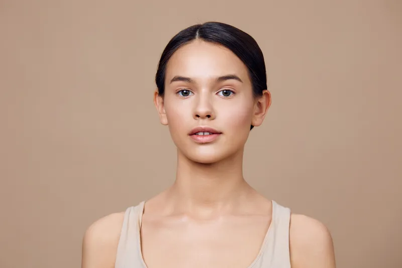 Natural beauty portrait of a young female model with a calm expression, wearing a simple beige top, against a smooth beige background