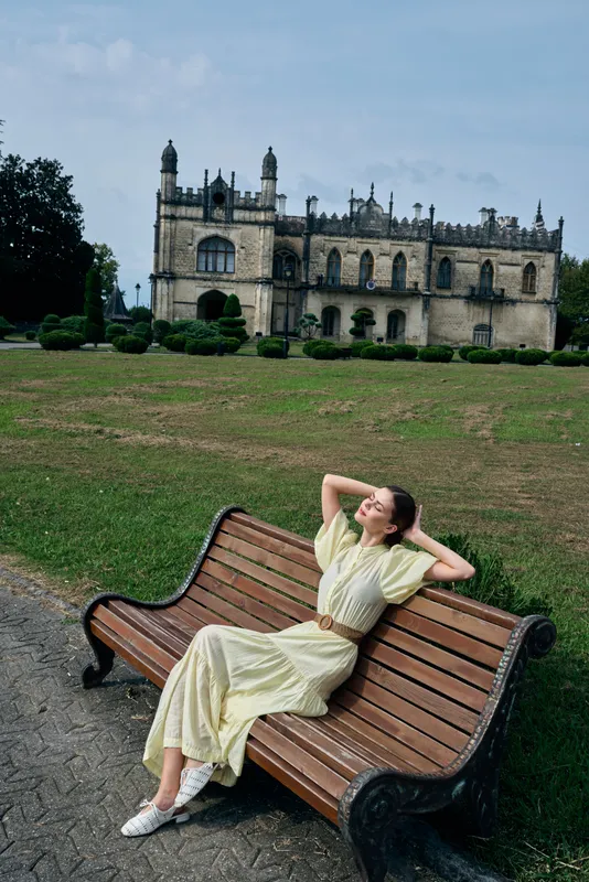 Woman relaxing on a wooden bench in a park with a historic castle in the distance, sunny day, casual dress, serene atmosphere and picturesque architecture and landscape