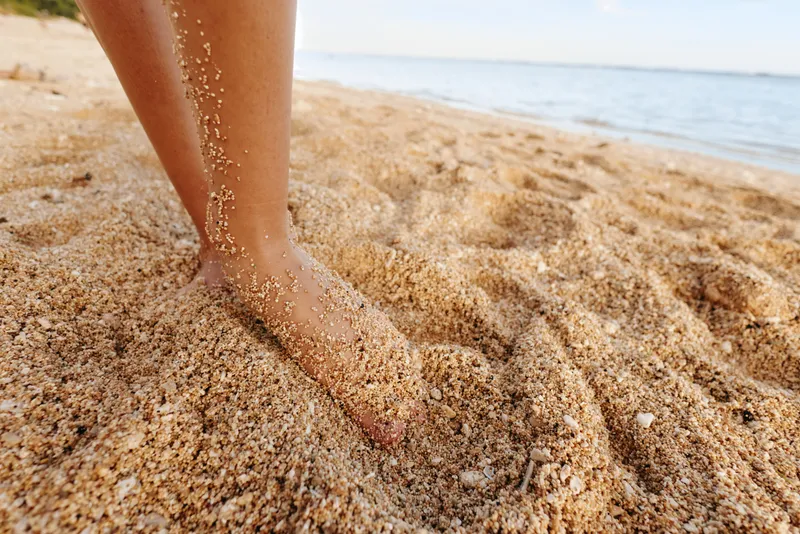 Beach sand, barefoot foot and legs walking along the ocean shoreline, summer sunlight highlights wet grains and footprints, outdoors calm coastal mood, closeup texture of seawater waves