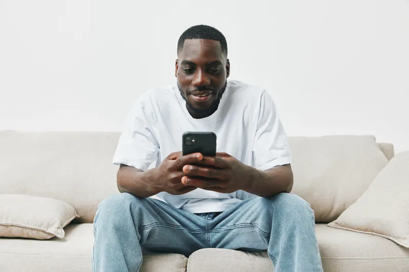 Happy black man sitting on a couch using a smartphone in a minimalistic living room, showcasing modern lifestyle and technology