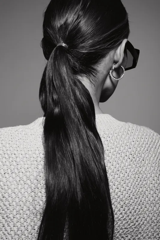 Back view of young Asian woman with long black hair tied in ponytail wearing black glasses and textured sweater against gray background, concept for hairstyle and fashion.