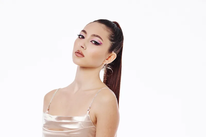 Young woman with sleek high ponytail wears glossy satin top, showcasing creative pink eye makeup on white background. Concept of beauty, fashion, and artistic style.