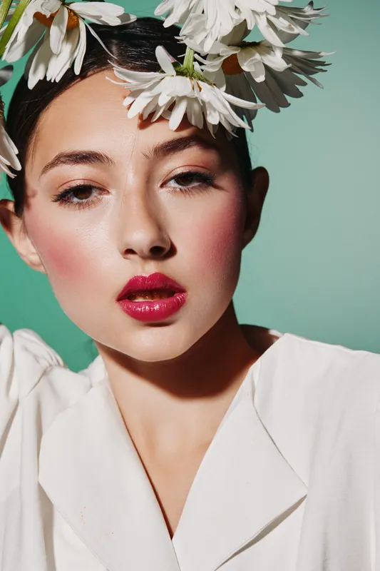 Closeup beauty portrait of young woman with glossy pink lips and rosy blush. Elegant daisies on her head. Bright fashion image with creative makeup on muted green background.