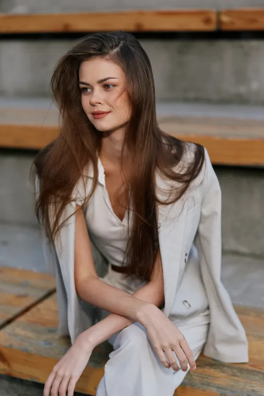 Young woman with long hair in light suit sitting on wooden steps, looking away thoughtfully with natural makeup and soft smile, fashion concept outdoor