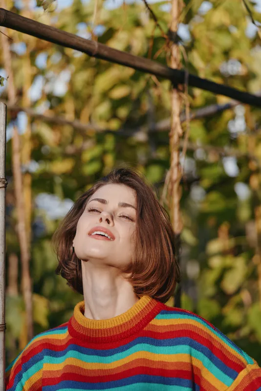 Woman portrait in a colorful rainbow sweater outdoors, sunlight on face, relaxed expression with closed eyes, short hair, natural background and warm golden light.