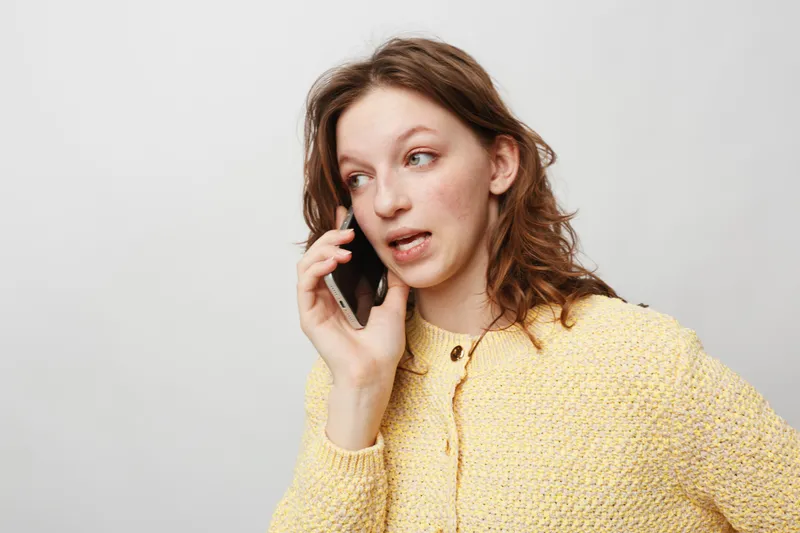 Young woman in yellow sweater talking on smartphone with concerned expression. Isolated on white background. Concept of communication, conversation, and emotion.