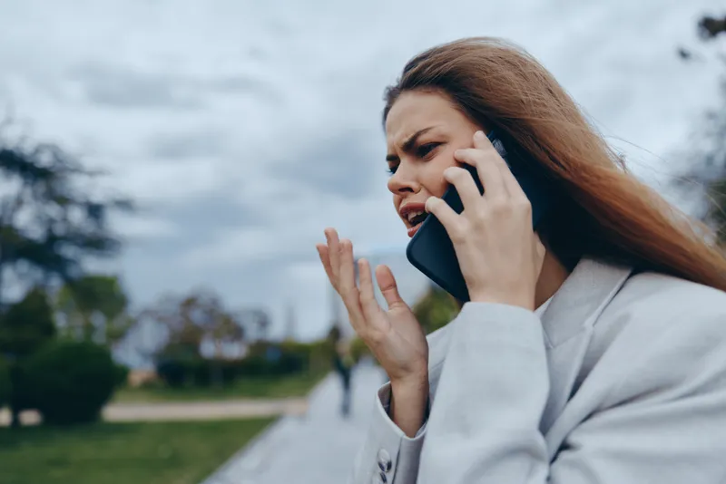 Business woman talking on smartphone, expressing frustration in an outdoor setting with overcast skies, conveying determination and professionalism.