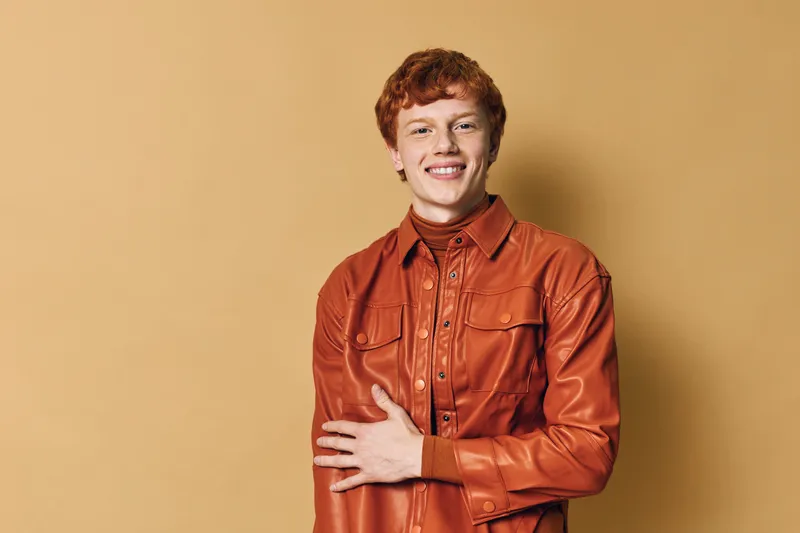 Young redhead man in orange leather jacket smiling with crossed arms against beige background. Casual clothing, hairstyle, confident, happy expression, studio portrait, isolated.