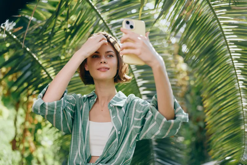 A young woman takes a selfie in a tropical garden, wearing a striped green shirt and white top, with sunlit palm leaves in the background.