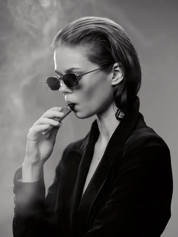 Stylish woman smoking cigarette in black and white portrait with sunglasses and dark outfit, exuding mystery and elegance.
