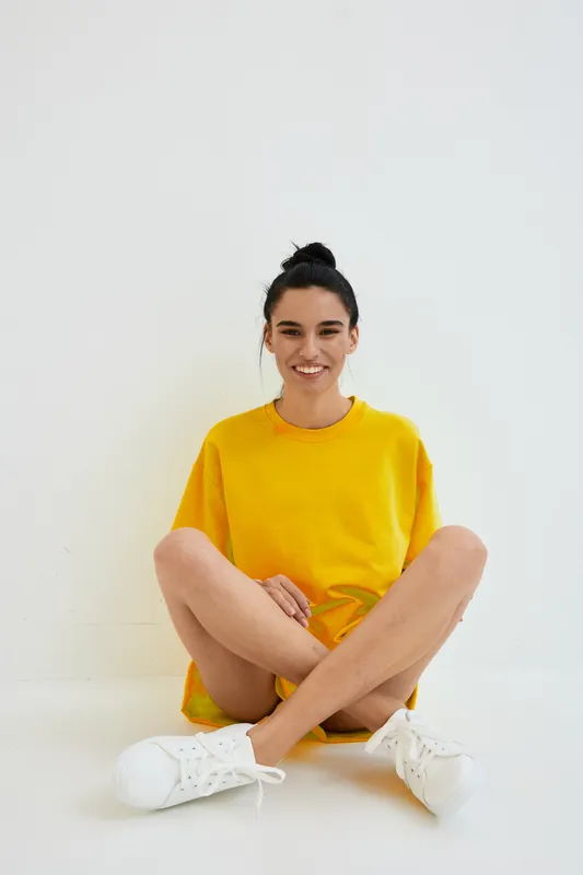 smiling woman in yellow oversized sweater sitting cross-legged on white floor, casual outfit, bright, cheerful mood, minimalistic background