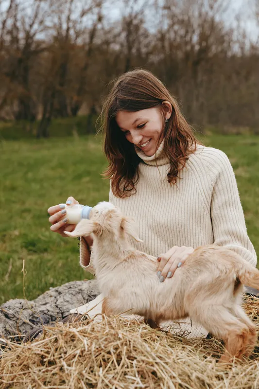 Smiling woman feeding a baby goat outdoors on a rustic farm. Soft sweater, straw, and green field create a wholesome lifestyle moment focused on care and animal companionship and care.