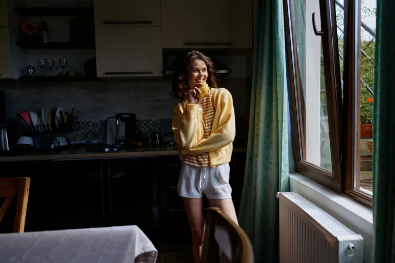 Young woman in a cozy kitchen wearing a yellow sweater and white shorts smiles softly by a sunlit window, feeling relaxed and casual as she stands near a table, gently touching her hair.