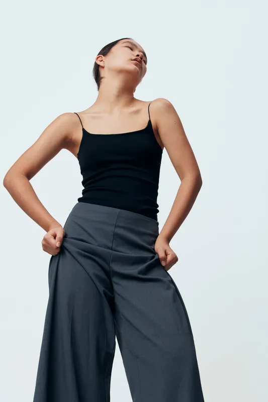 Woman posing in black tank top and wide leg gray pants looking away with hands in pockets against plain background. Fashion model with minimal style and confident posture in studio.