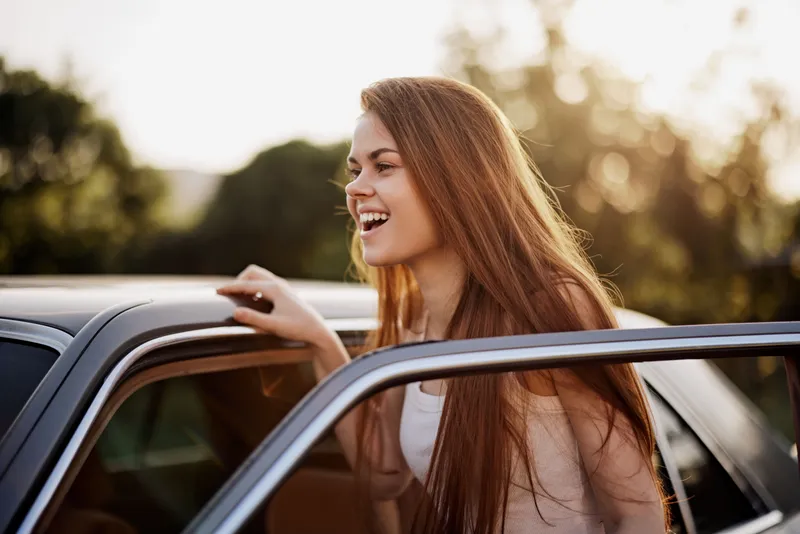 Young woman with long hair smiling joyfully while leaning out of a car during sunset, showcasing a carefree summer vibe and a sense of adventure.
