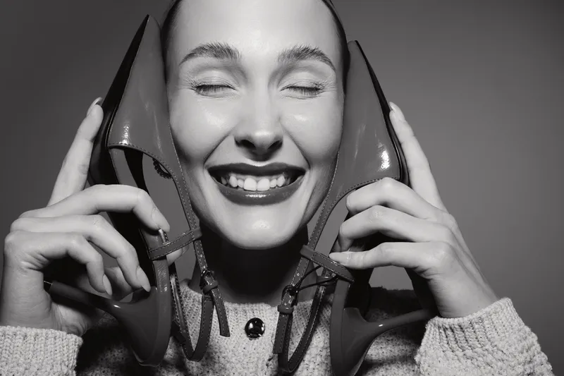Young Caucasian woman with fair skin smiles joyfully holding two high heel shoes near her face in black and white studio portrait. Concept of happiness and fashion accessories.