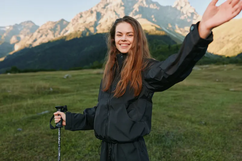 A cheerful outdoor adventurer stands in a wide meadow with towering mountains in the background. She smiles and raises one arm, holding a trekking pole, exuding energy and optimism.
