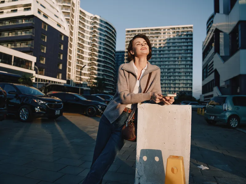 A stylish urban professional stands outdoors in a modern office district, looking up with a smile while checking her phone, conveying optimism and confidence in a dynamic city environment.