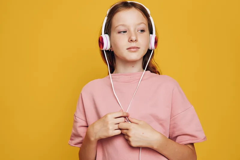 Teenage girl in pink t-shirt and white headphones listens music looking aside on yellow background. Studio portrait with casual style and relaxed mood.