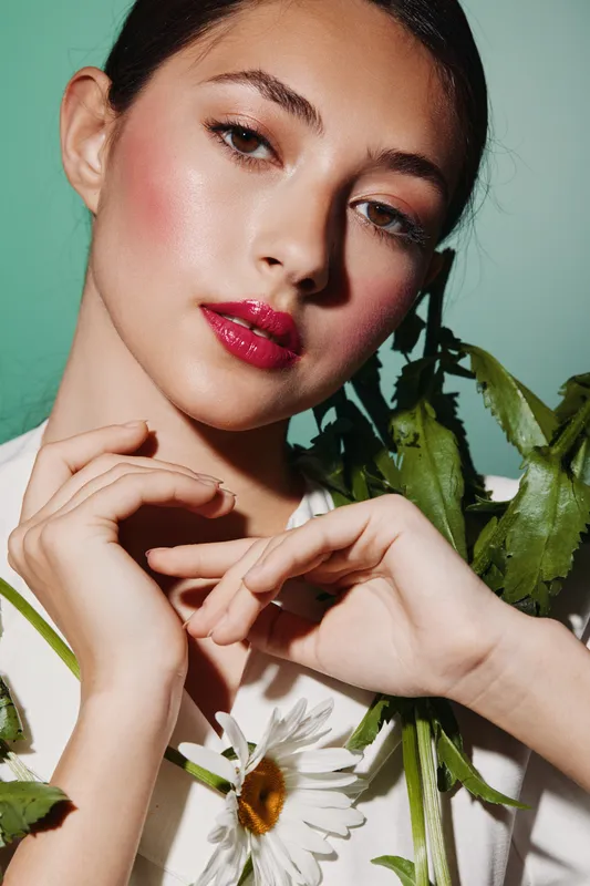 Close-up portrait of a young woman with glossy pink lips and dewy skin holding white daisy flowers against pastel green background, vibrant beauty concept blending fashion and nature.