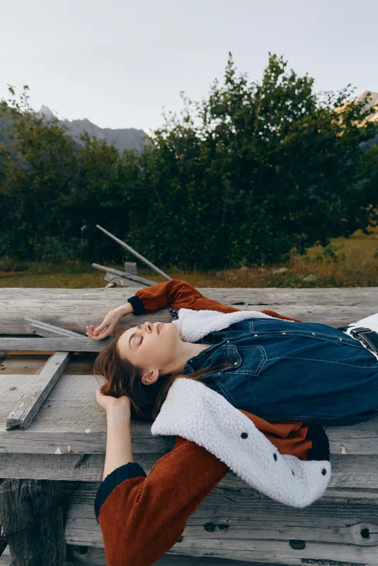 Woman portrait relax outdoors in nature on wood dock, wearing jacket and layered clothing for leisure, eyes closed in a peaceful casual fashion scene and calm lifestyle moment.