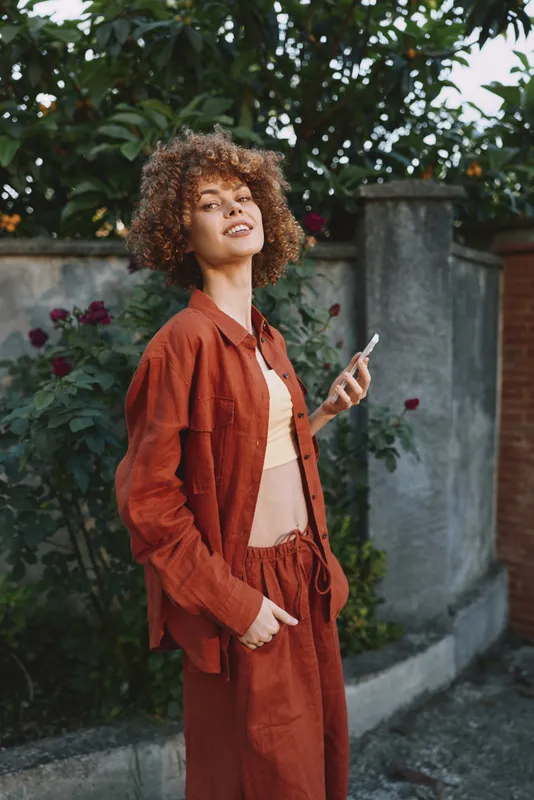 Young woman with curly hair stands outdoors in a rust outfit, smiling as she holds a smartphone, posing casually in a sunlit garden with bright flowers and a stone wall behind, with a soft breeze